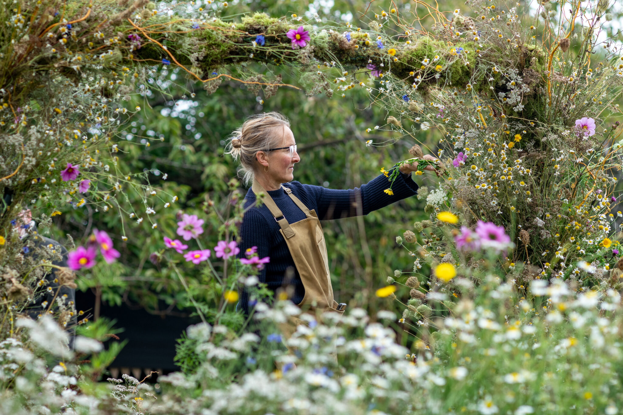Close up shot of gardener with long fair hair and an apron on, tending to a wild flower archway and meadow. 