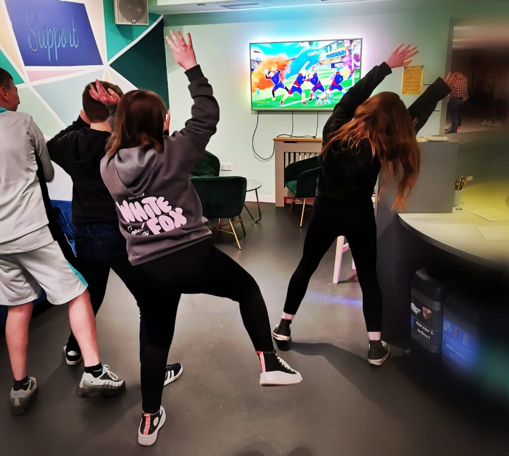 Young people dancing with their hands in the air, whilst watching an interactive dance game on screen.