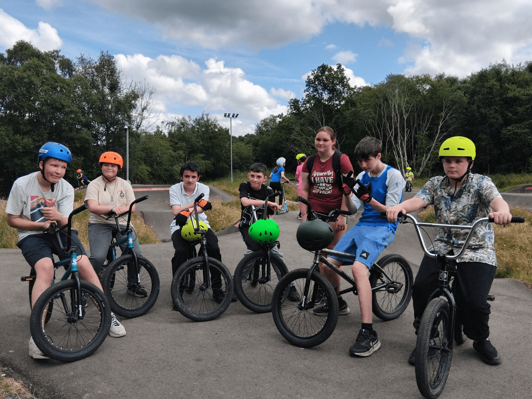 Group of young people with BMX bikes gathered at an outdoor skate park in daytime, wearing helmets in bright colors. Trees and ramps are visible in the background under a partly cloudy sky.