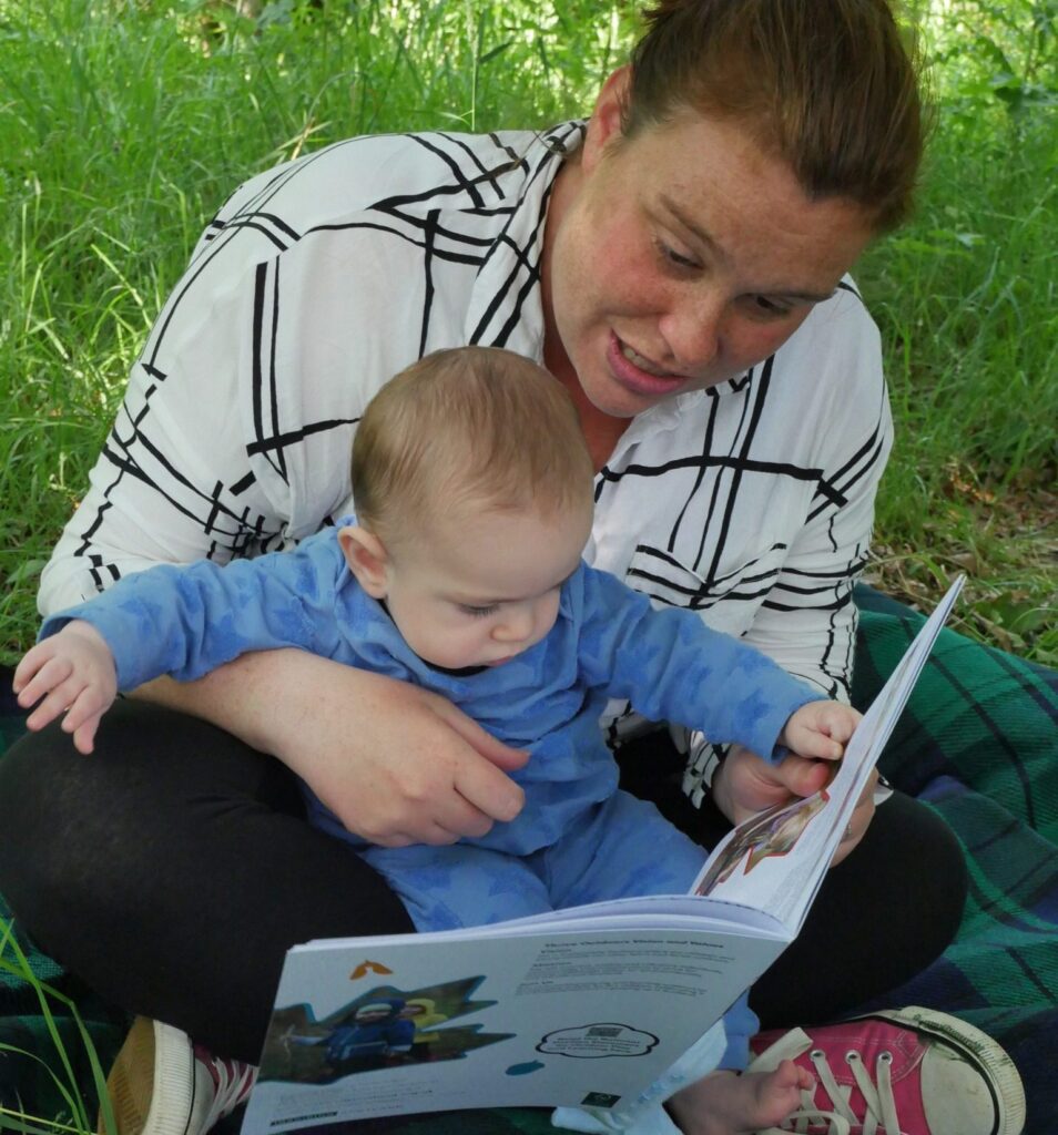 Adult and baby sitting on grass and reading a story book.