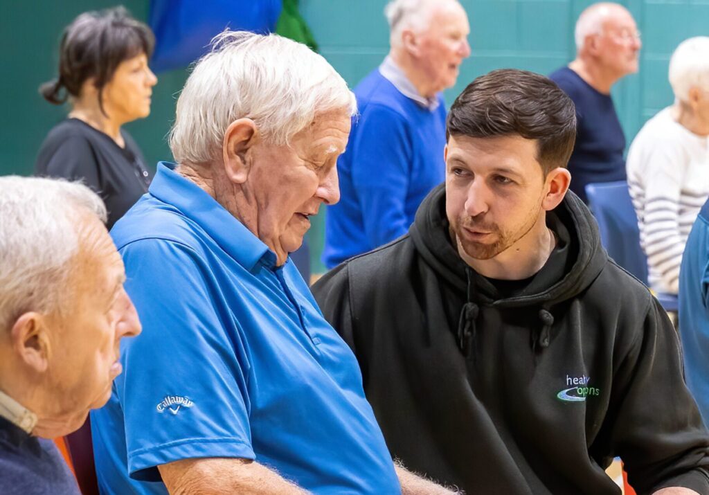 A Healthy Options staff member in a crouched position and talking with people seated in a group during an indoor exercise or wellbeing session.