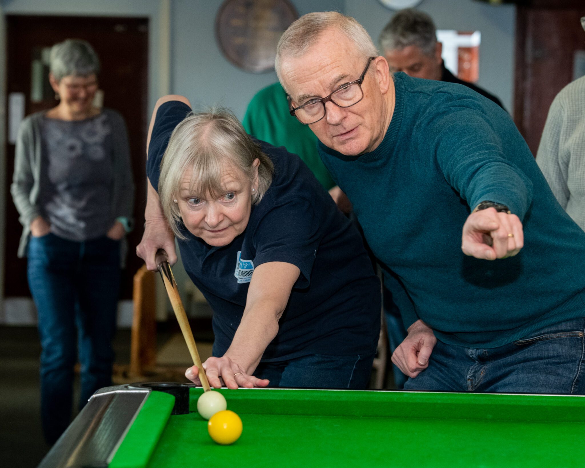 Two people standing at a pool table, with one person guiding another as they take a shot.