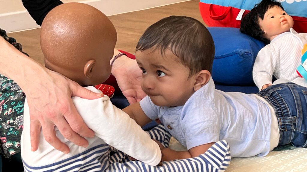 Baby lying on it's tummy interacting and making eye contact with a baby doll.