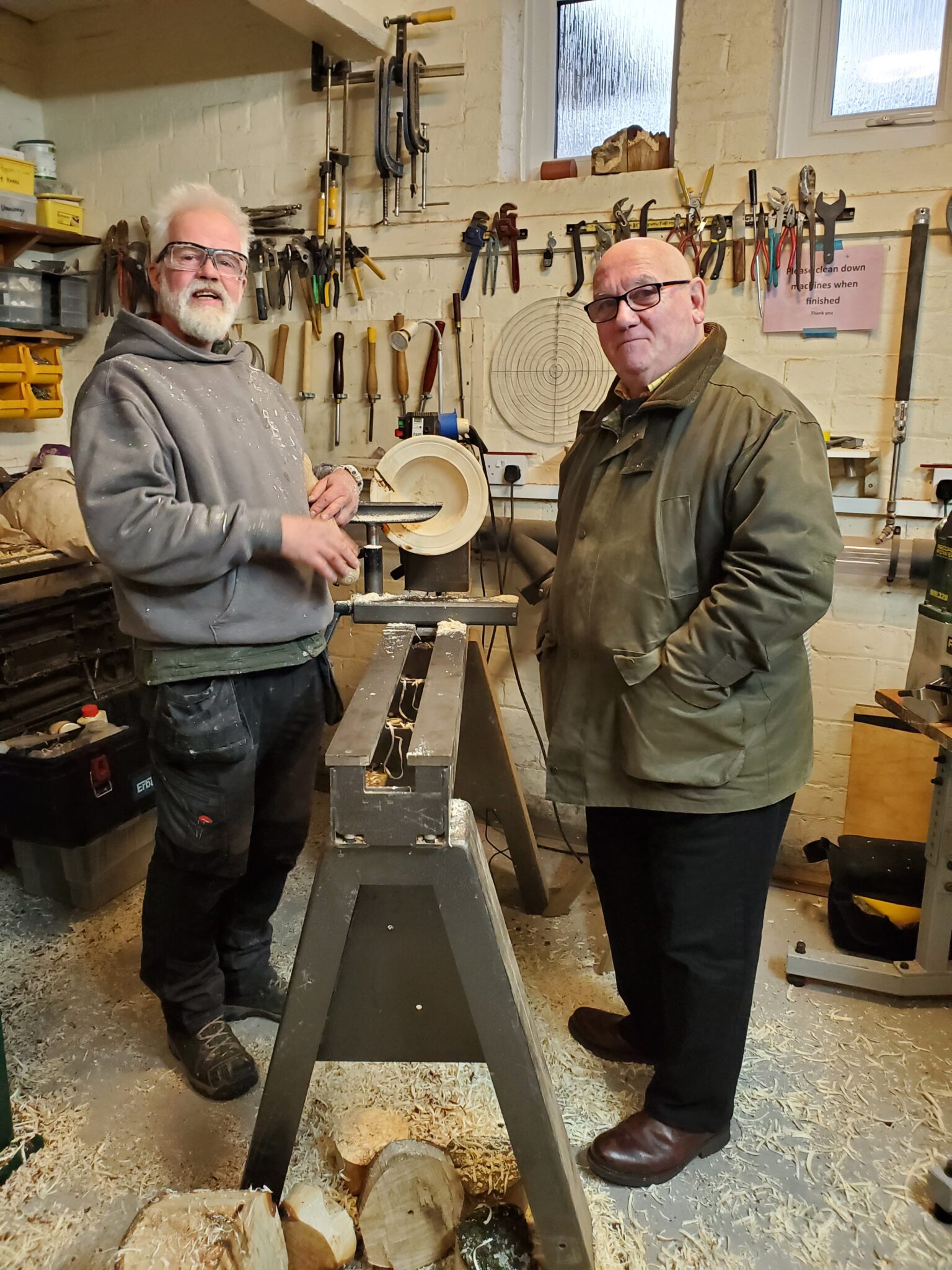 Two men standing in a Men's shed with tools on the wall behind them.